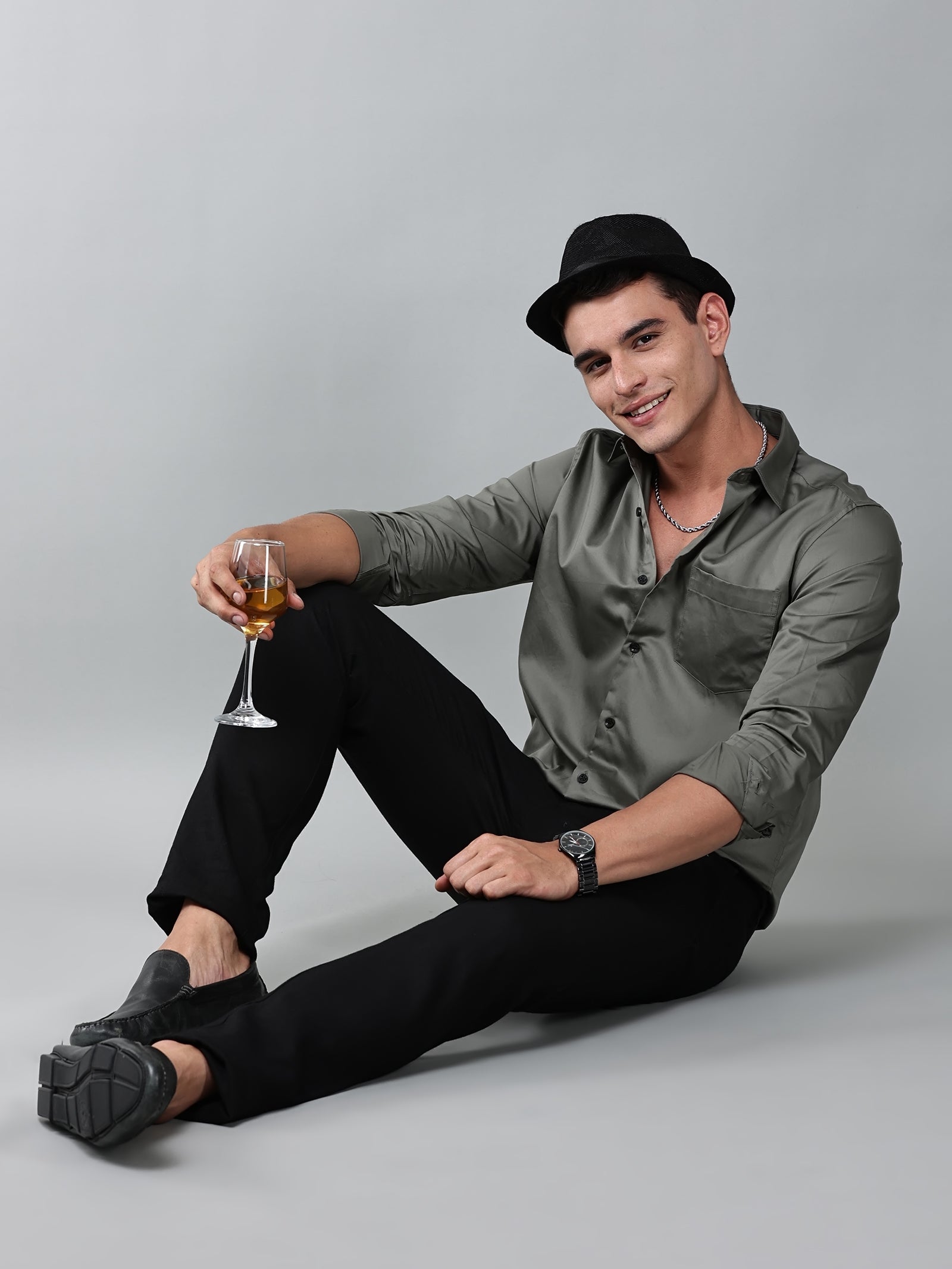a model wearing a luxe satin olive green shirt, black trousers, black loafers and a black hat with a glass in one hand