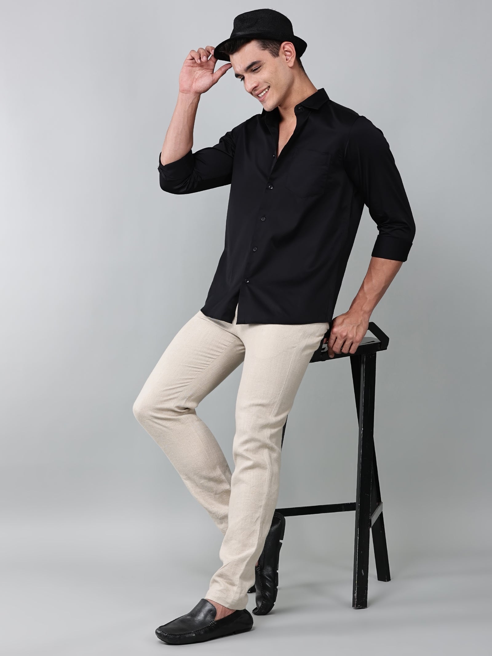 a model wearing a luxe satin black shirt with beige trousers, black loafers and a black hat