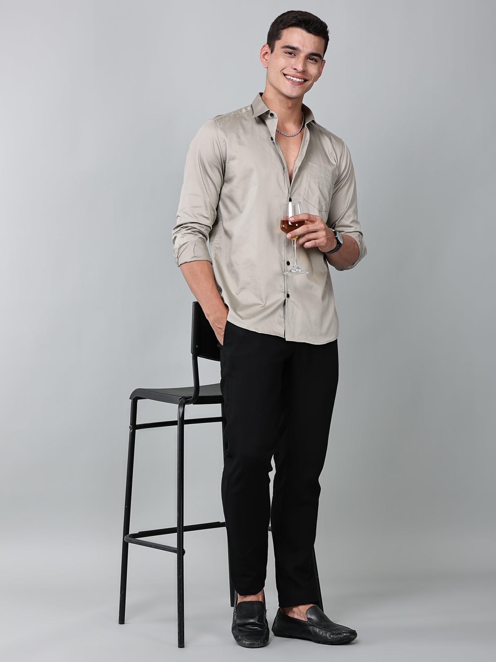 a model wearing a luxe satin ivory stone shirt with black trousers and black loafers and holding a glass in one hand