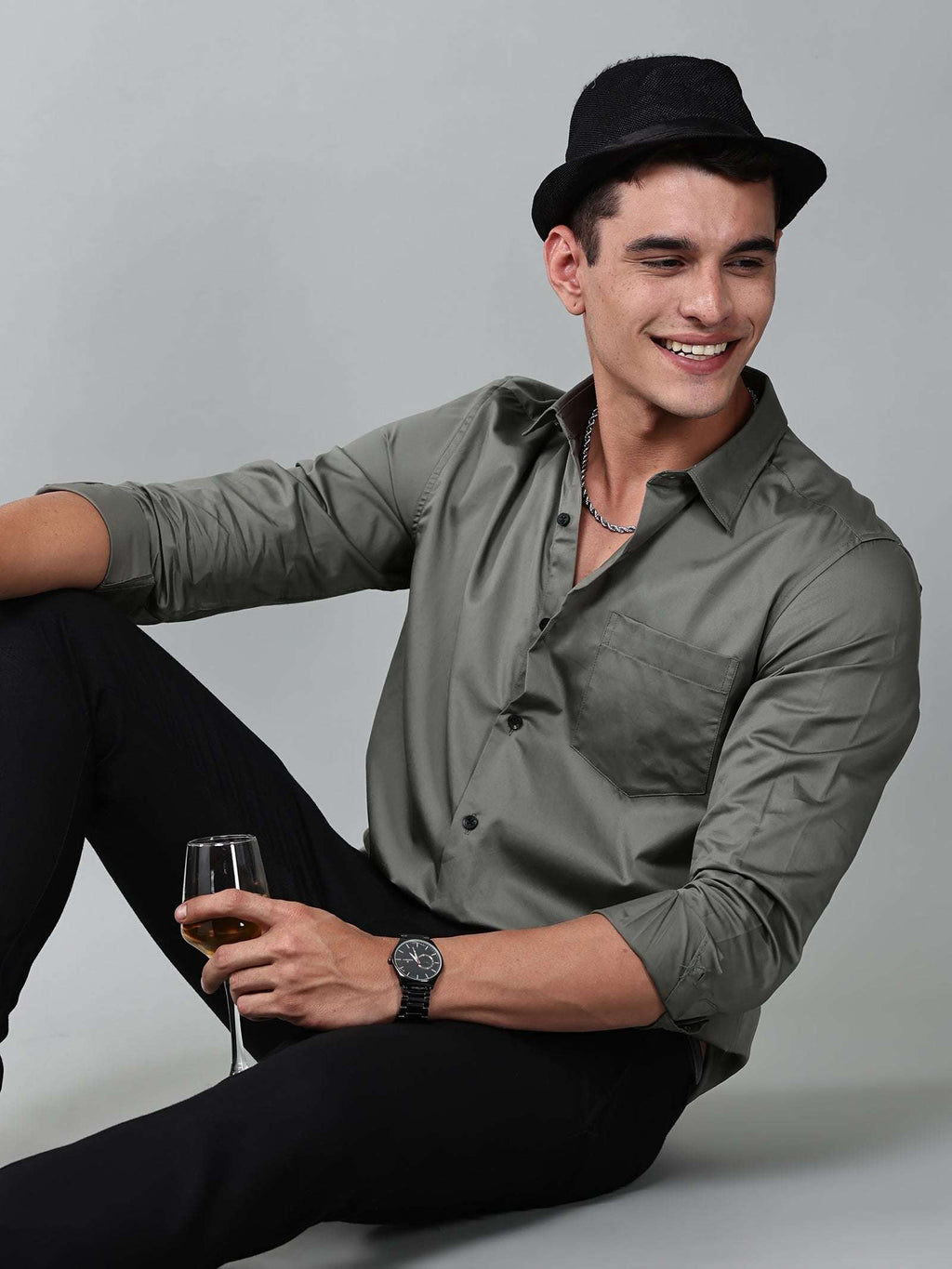 a model wearing a luxe satin olive green shirt with a black hat holding a glass in one hand